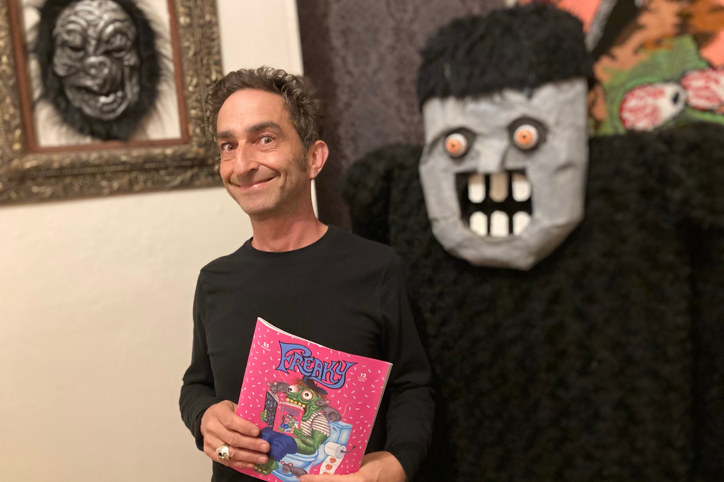 A man posing with an impish expression holding a pink magazine called Freaky, standing in front of a paper mache monster.