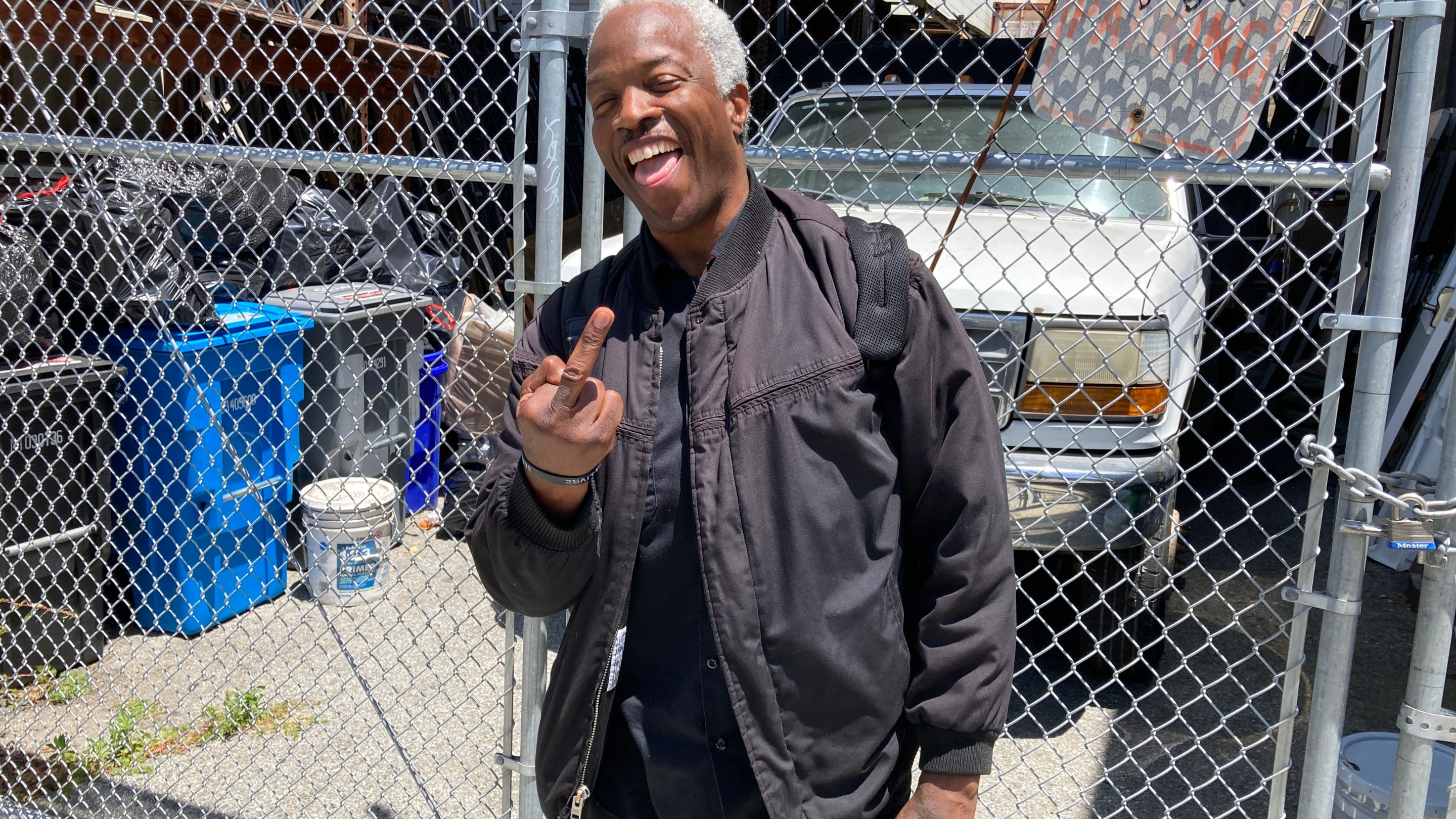 A man in a black jacket is flipping the bird and smiling while standing by chain link fence.