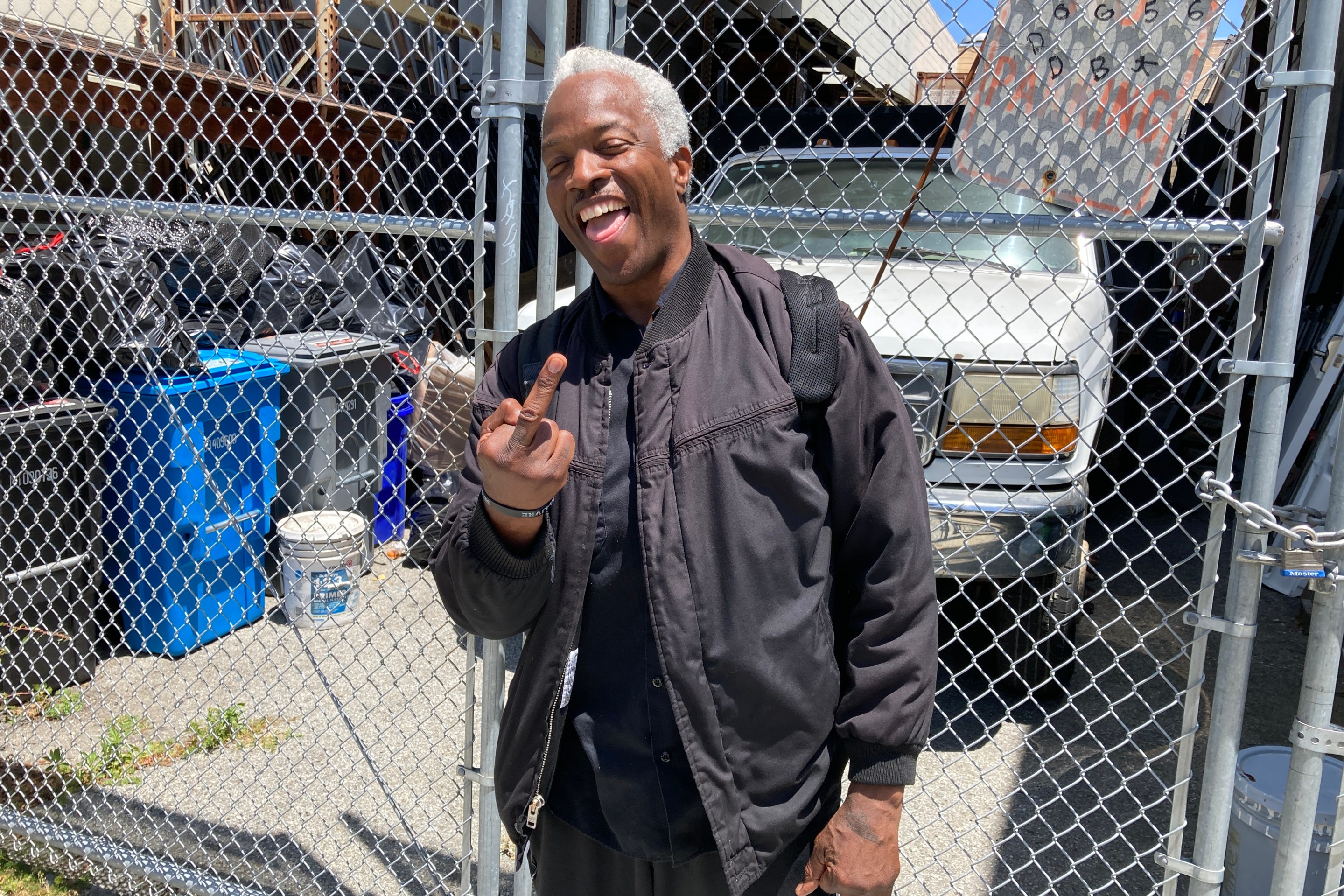 A man in a black jacket is flipping the bird and smiling while standing by chain link fence.