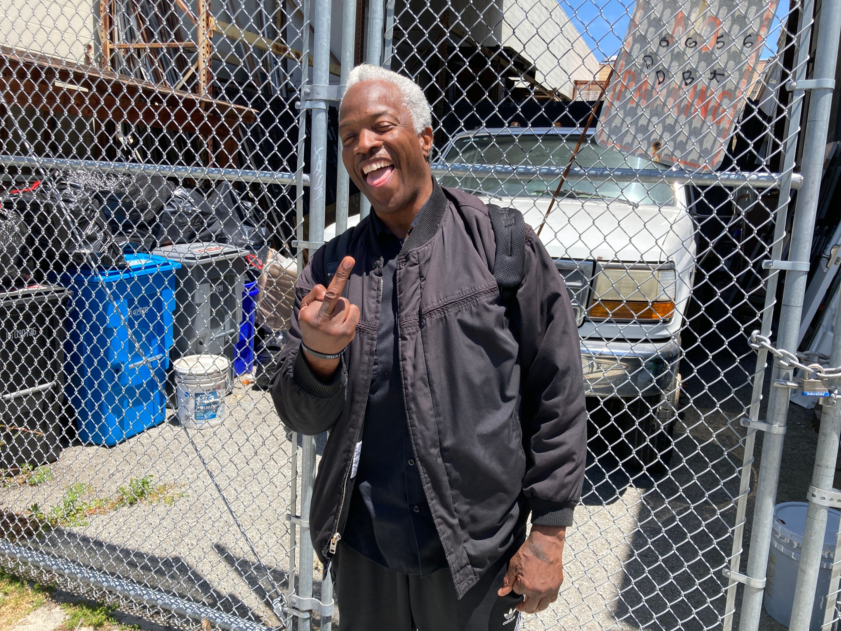 A man in a black jacket is flipping the bird and smiling while standing by chain link fence.