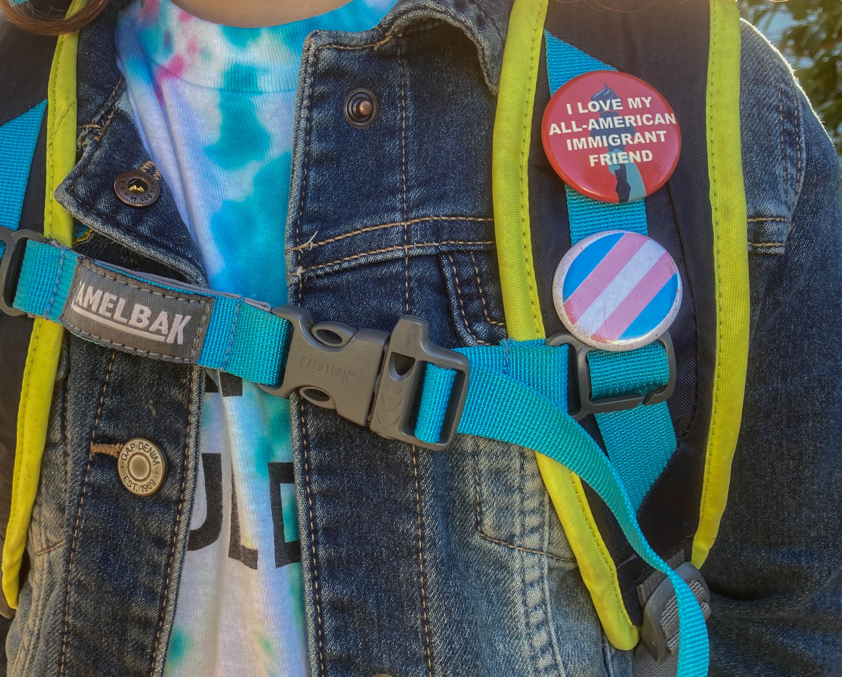 Closeup photo of a child's backpack with two pins: One reads I Love My All-American Immigrant Friend; the other is a trans pride flag.