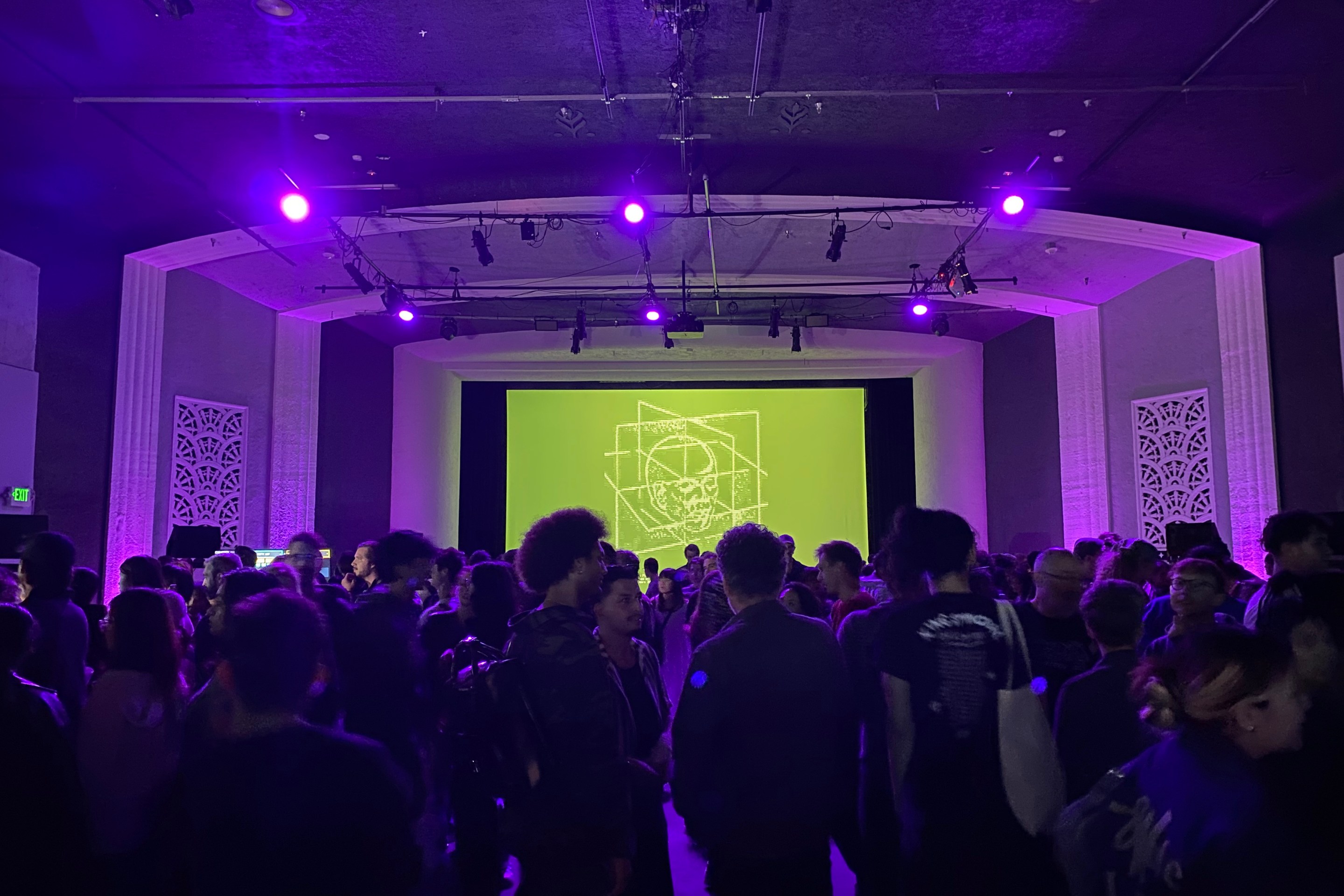 A room lit with purple lights is packed with people, backlit by a projected abstract image of a human head with mathematical line art on a green background.