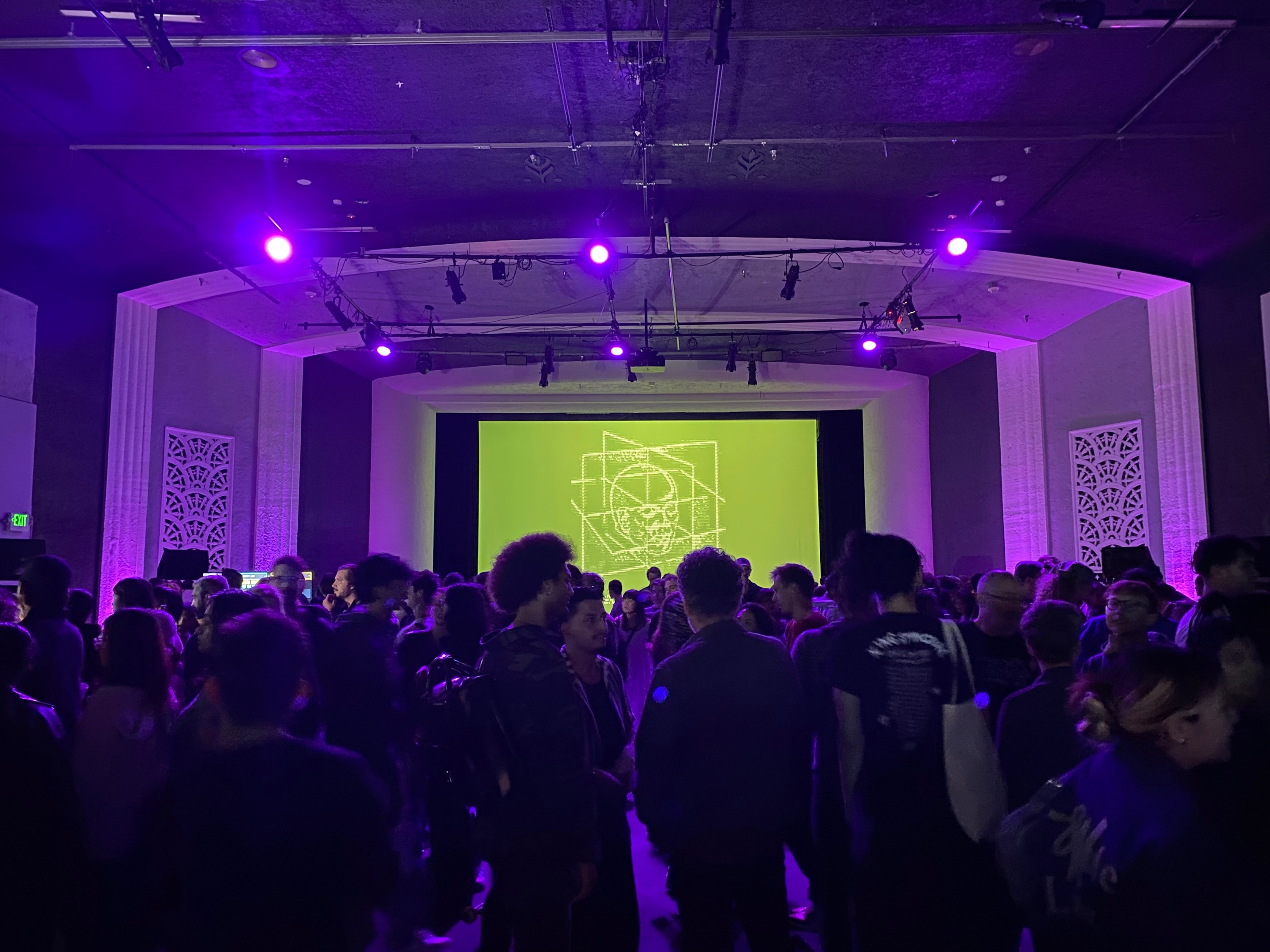 A room lit with purple lights is packed with people, backlit by a projected abstract image of a human head with mathematical line art on a green background.