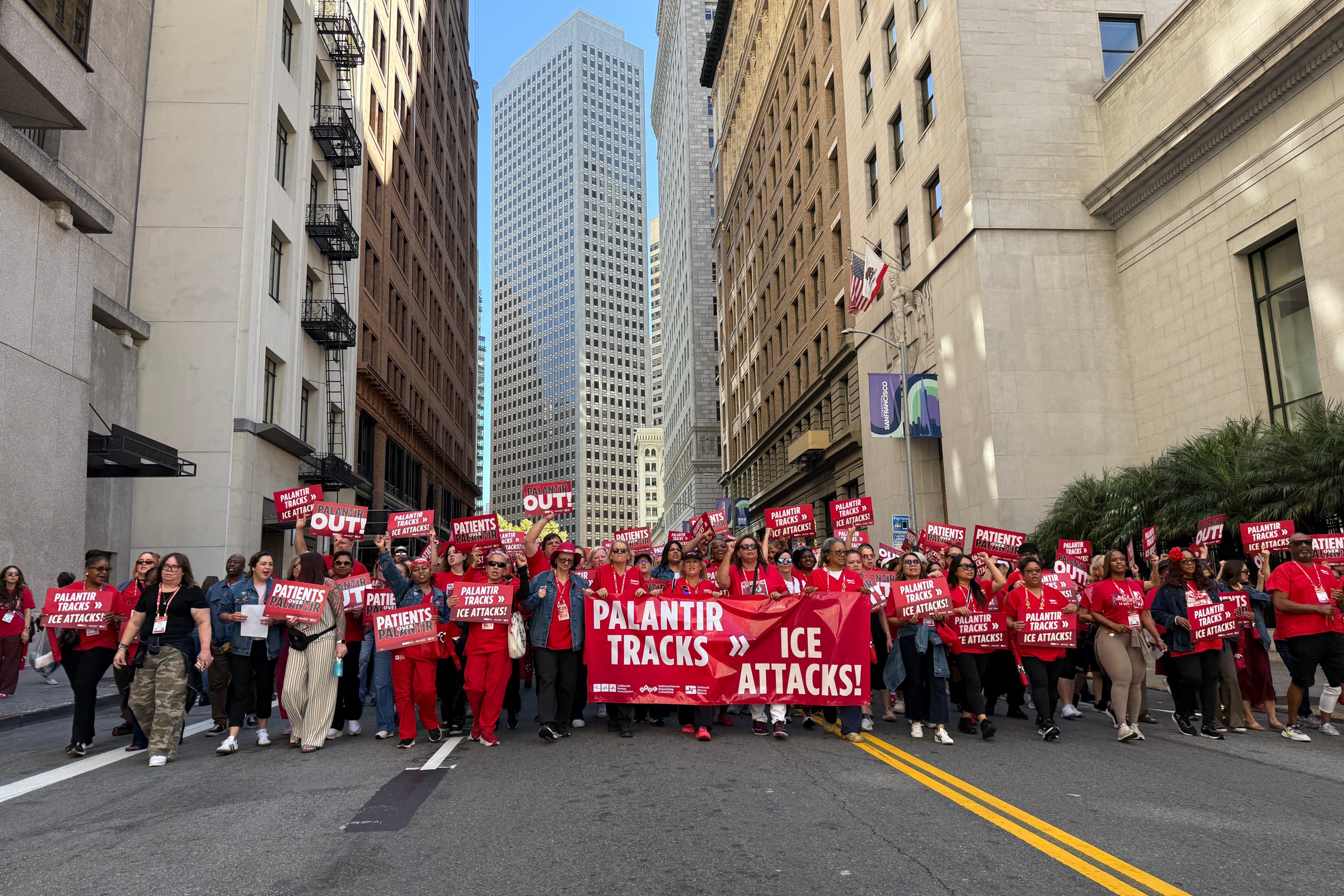 Large crowd of protesters holding red signs marching down city street against Palantir and ICE.