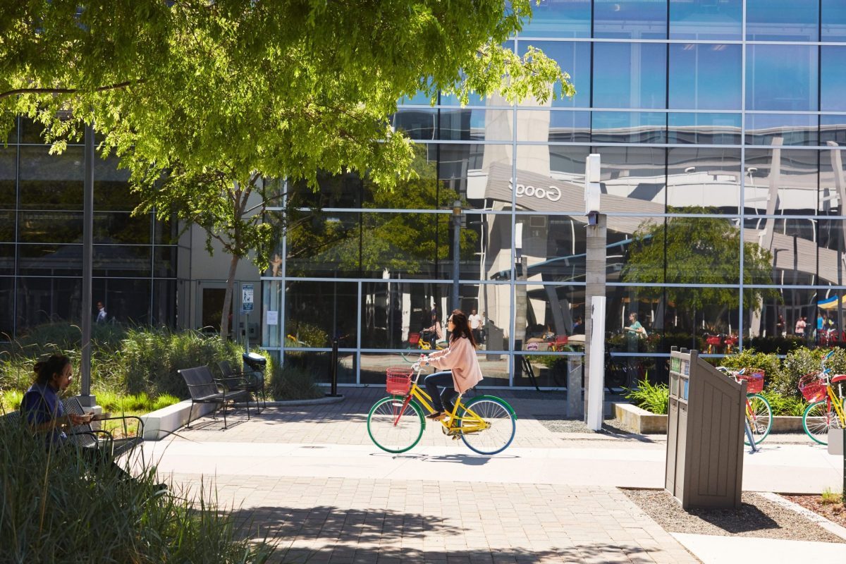 Woman riding a yellow bicycle in a modern urban plaza with green trees and contemporary buildings