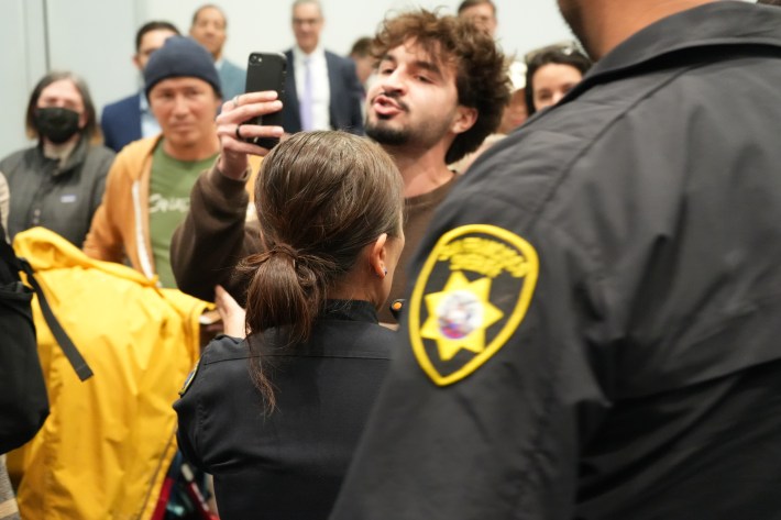 Police officers interact with a man recording on smartphone amid gathered crowd indoors.