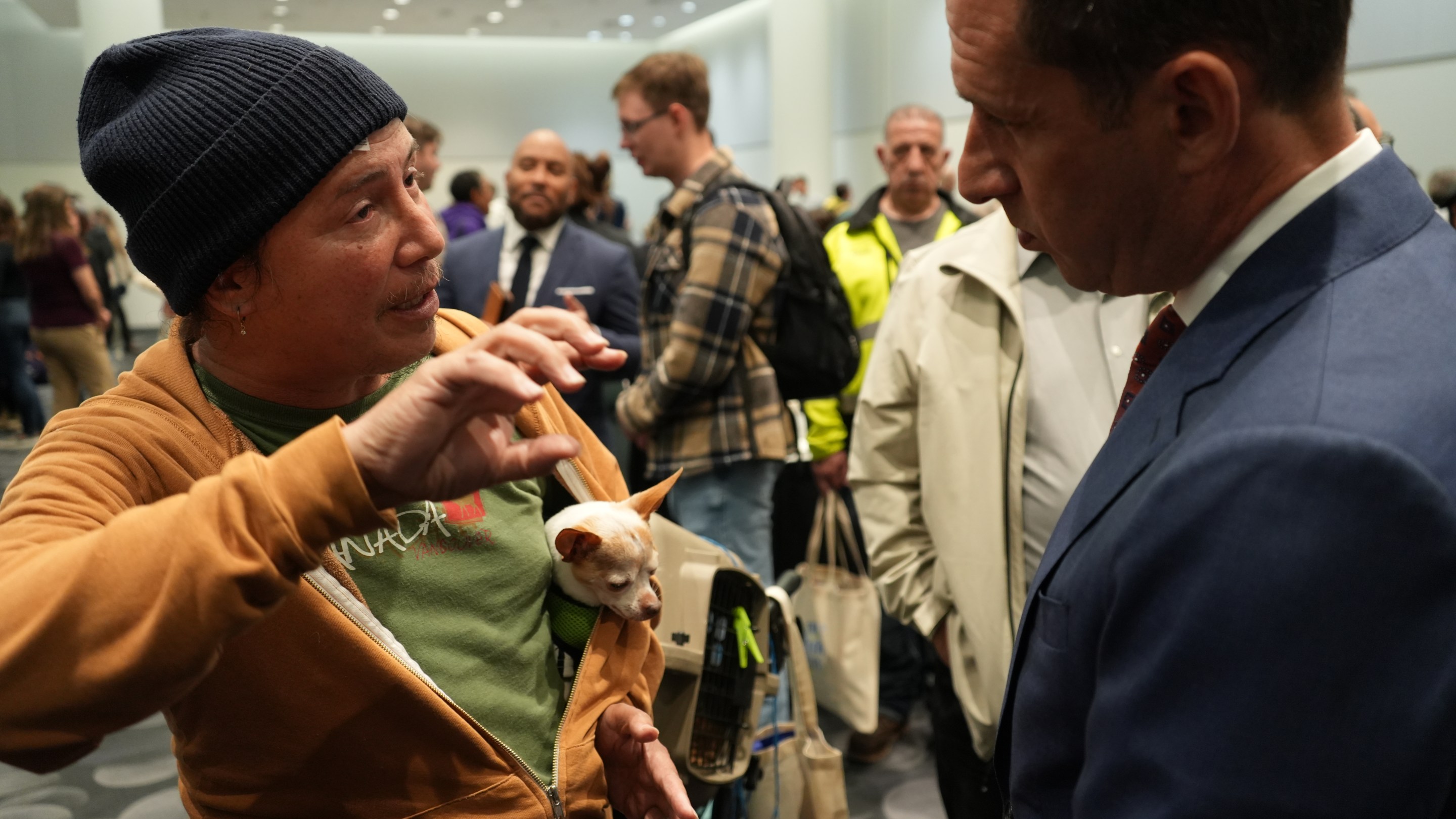 Man in beanie holds meerkat while speaking with man in blue suit at indoor event