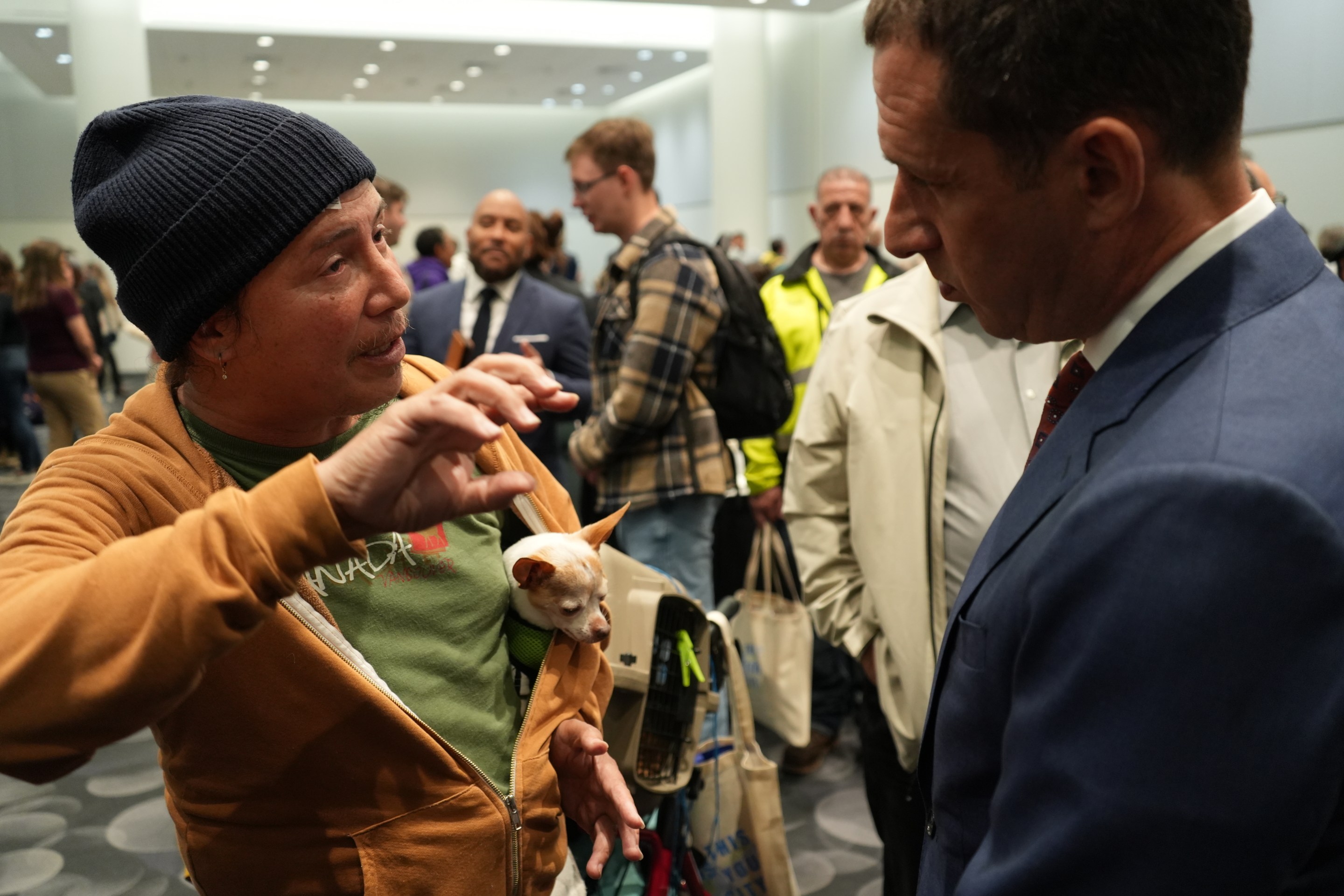 Man in beanie holds meerkat while speaking with man in blue suit at indoor event