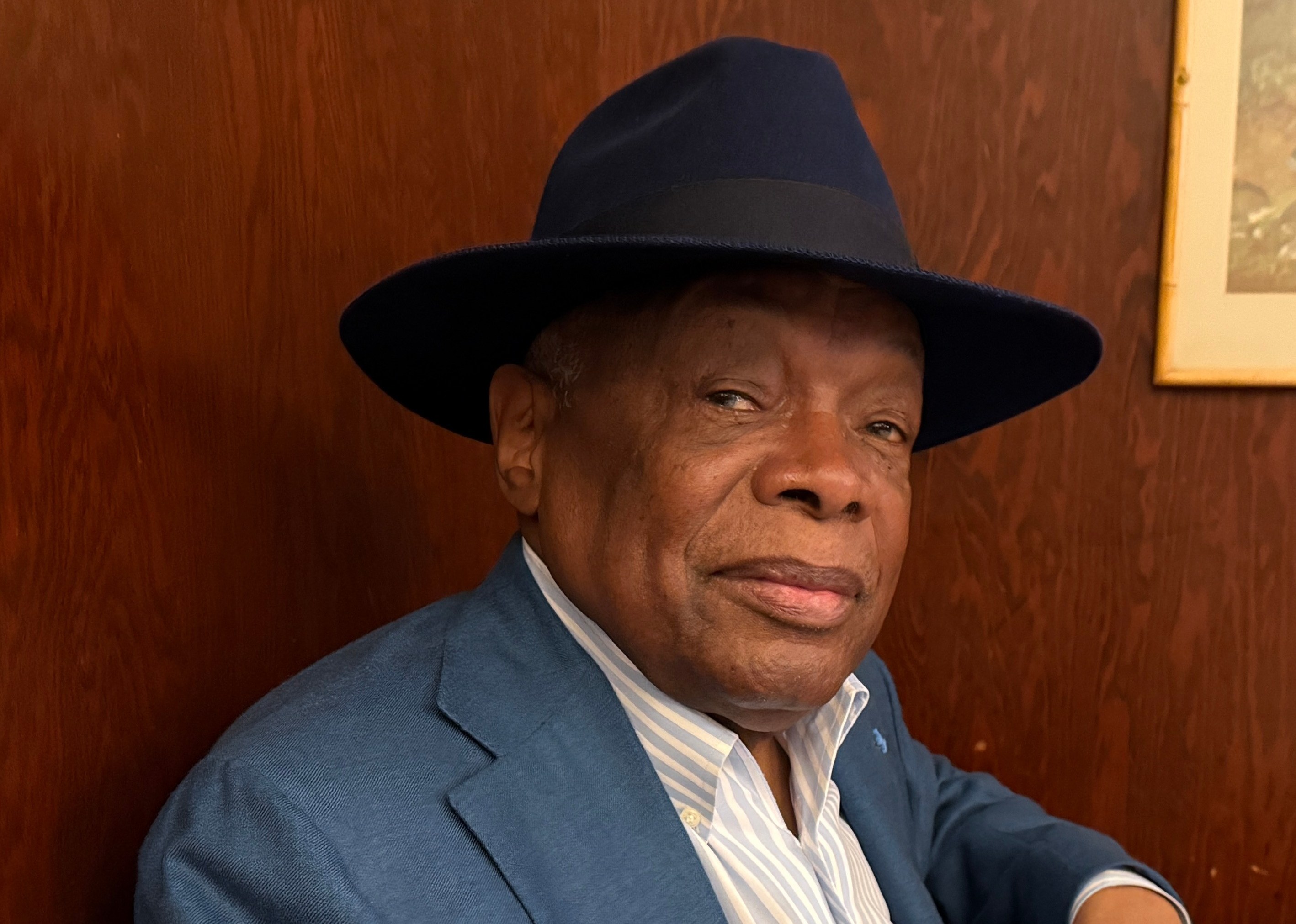 Distinguished man in blue suit and fedora hat seated at desk indoors.