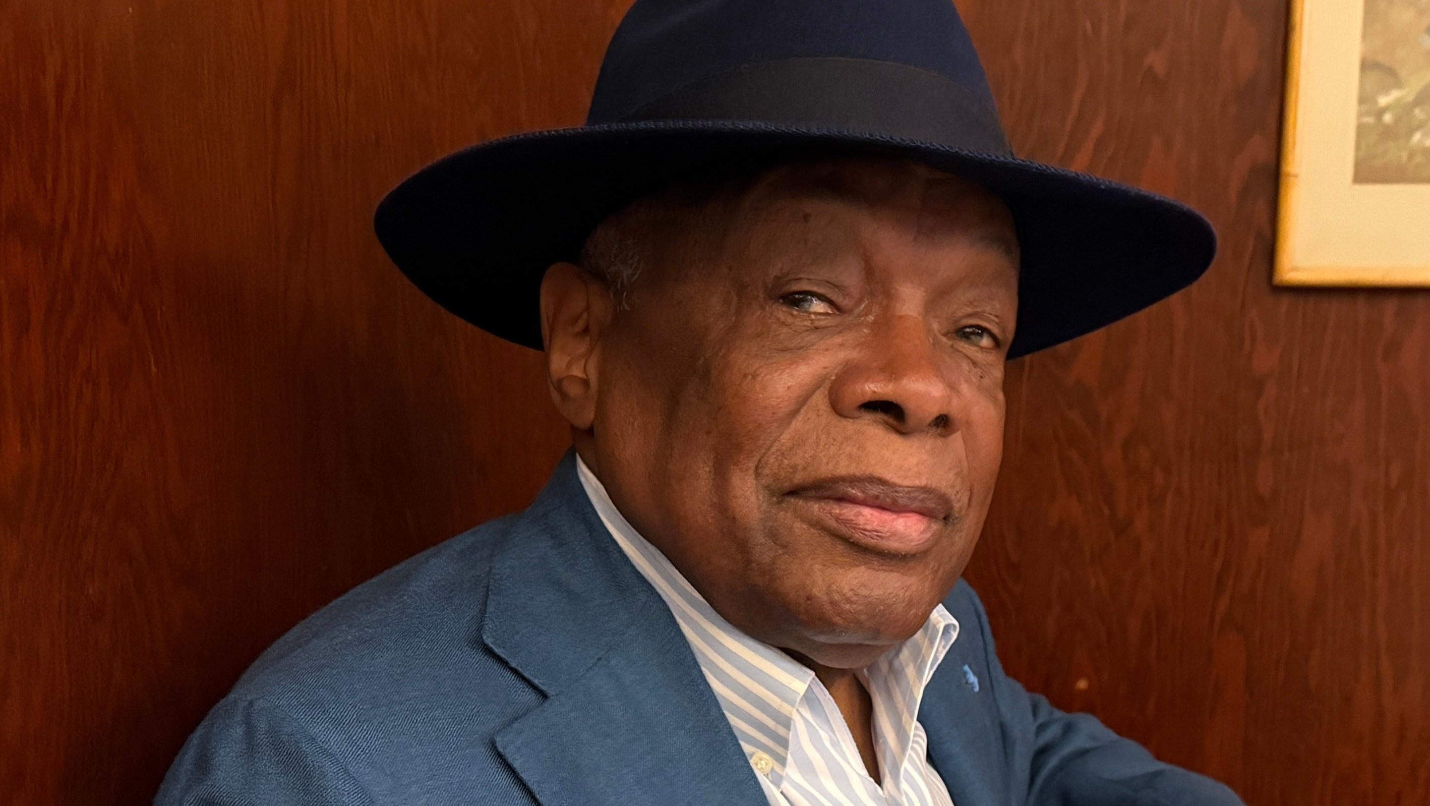 Distinguished man in blue suit and fedora hat seated at desk indoors.
