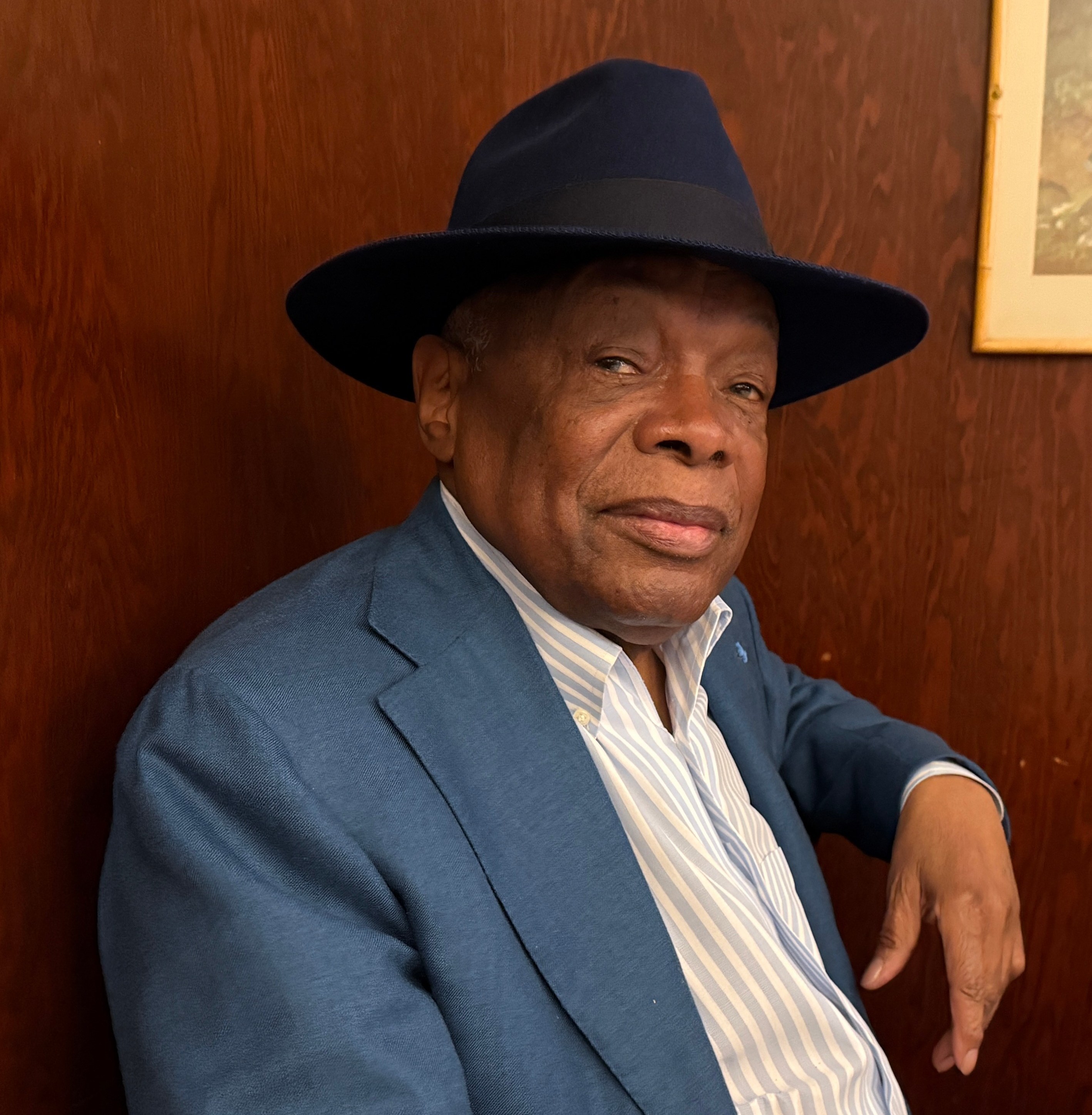 Distinguished man in blue suit and fedora hat seated at desk indoors.