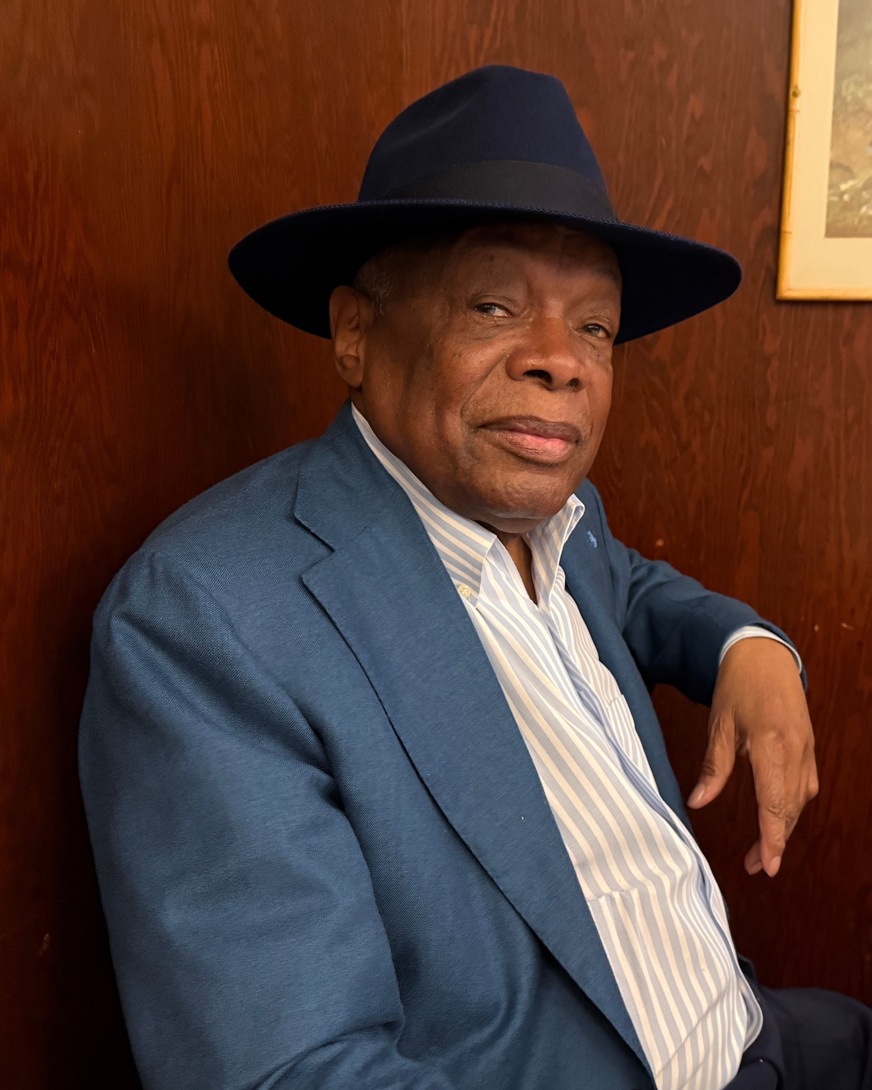 Distinguished man in blue suit and fedora hat seated at desk indoors.
