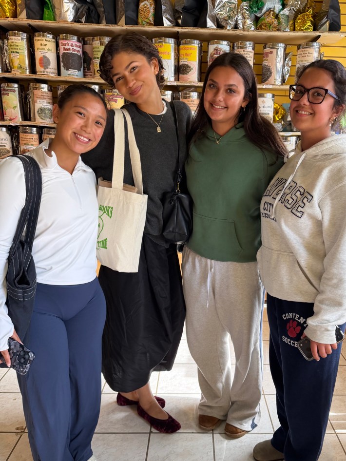Four young women smiling together in a bookstore with shelves of books behind them.
